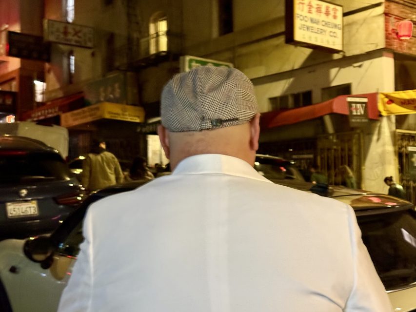 A person in a white jacket and checkered cap stands on a busy street at night, with illuminated signs and buildings in the background.