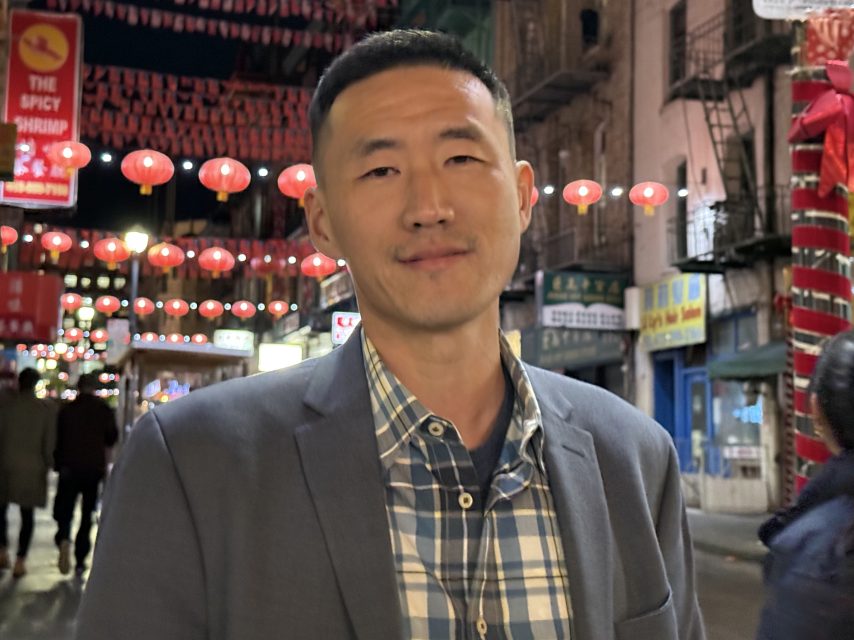 A person in a checkered shirt and blazer stands on a street decorated with red lanterns at night.