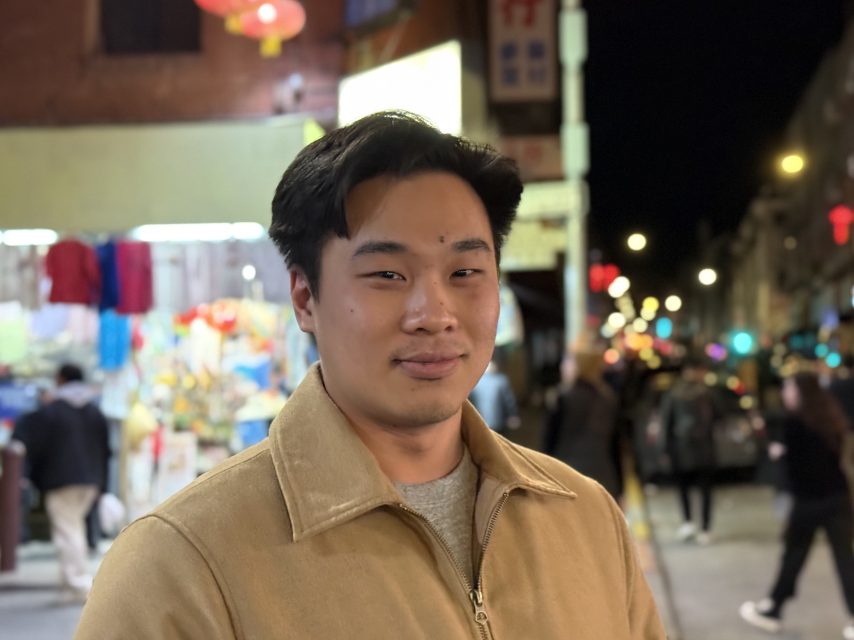 A person in a tan jacket stands on a bustling city street at night, with lit storefronts and blurred pedestrians in the background.