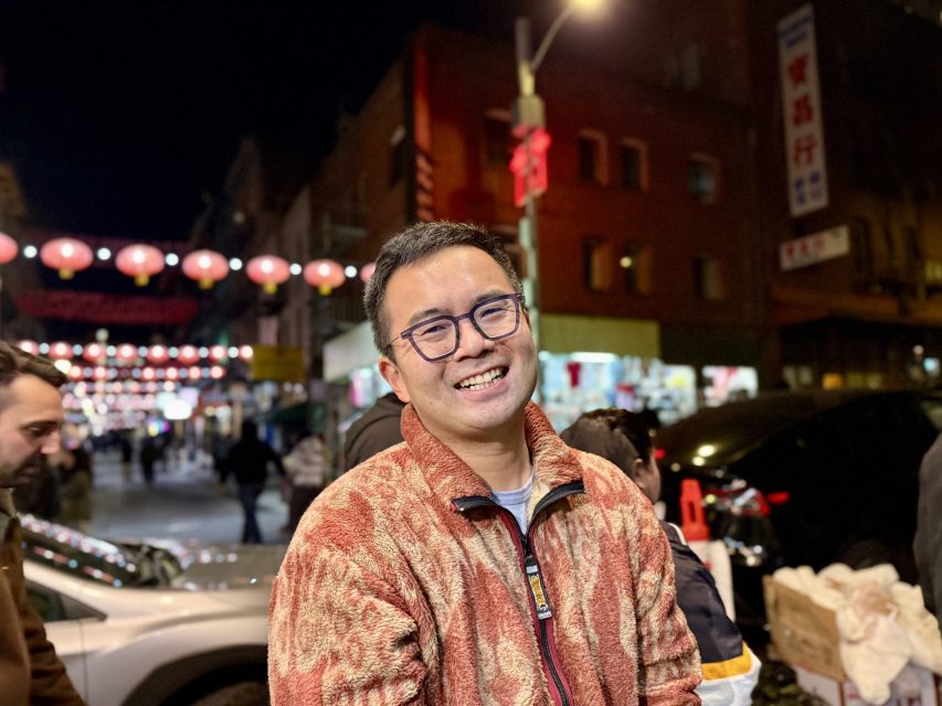 A person wearing glasses and a patterned jacket smiles while standing outdoors at night in a bustling street with decorative lanterns.