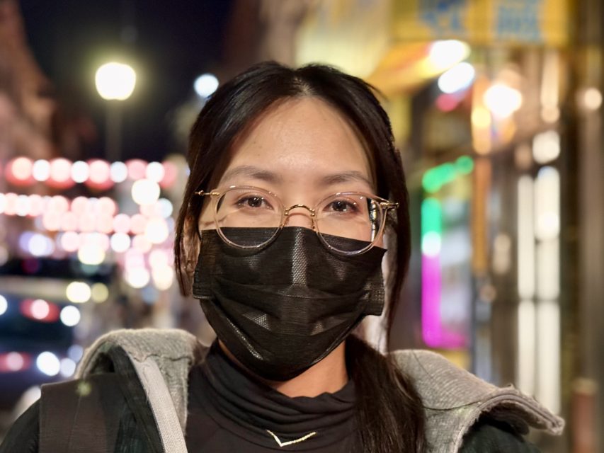 A person wearing glasses and a black face mask stands outdoors at night, with blurred lights and colorful signs in the background.