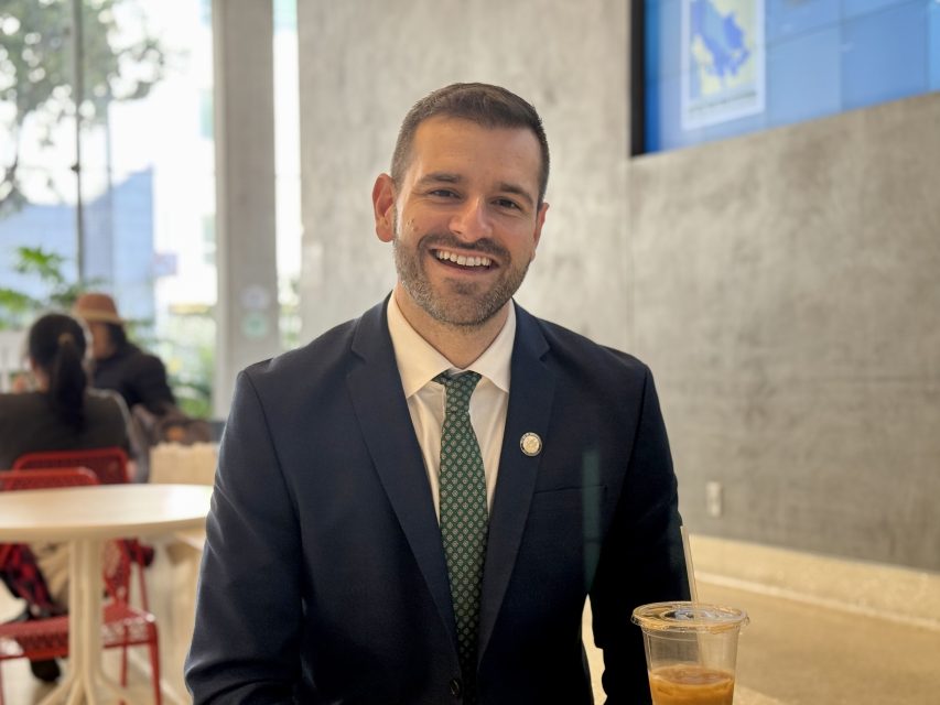 Danny Sauter in a suit with a green tie sits at a table with an iced coffee, smiling in a modern café setting.