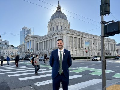 Danny Sauter in a suit stands smiling at a crosswalk in front of a large, historic building with a dome. Pedestrians are crossing the street.