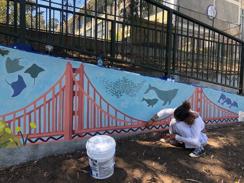 An artist paints a mural featuring a red bridge, sea creatures, and sky on a wall. A paint bucket and tools are on the ground nearby.