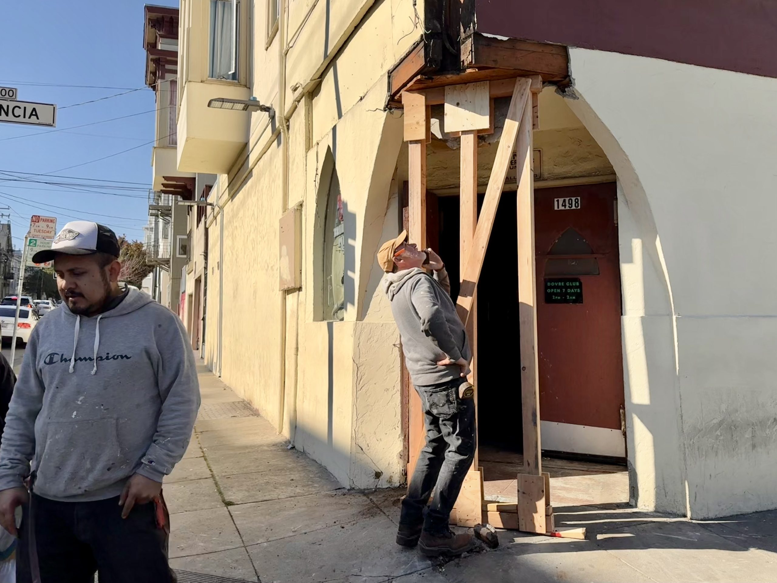 Man inspects wooden support structure outside a corner building on a sunny day; another man walks by on the sidewalk.