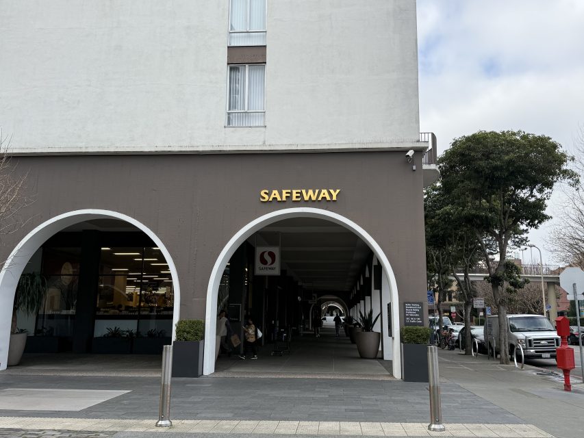 Entrance of a Safeway store with an arched facade, large illuminated sign, and a view of the interior. Trees and a sidewalk are visible in the foreground.
