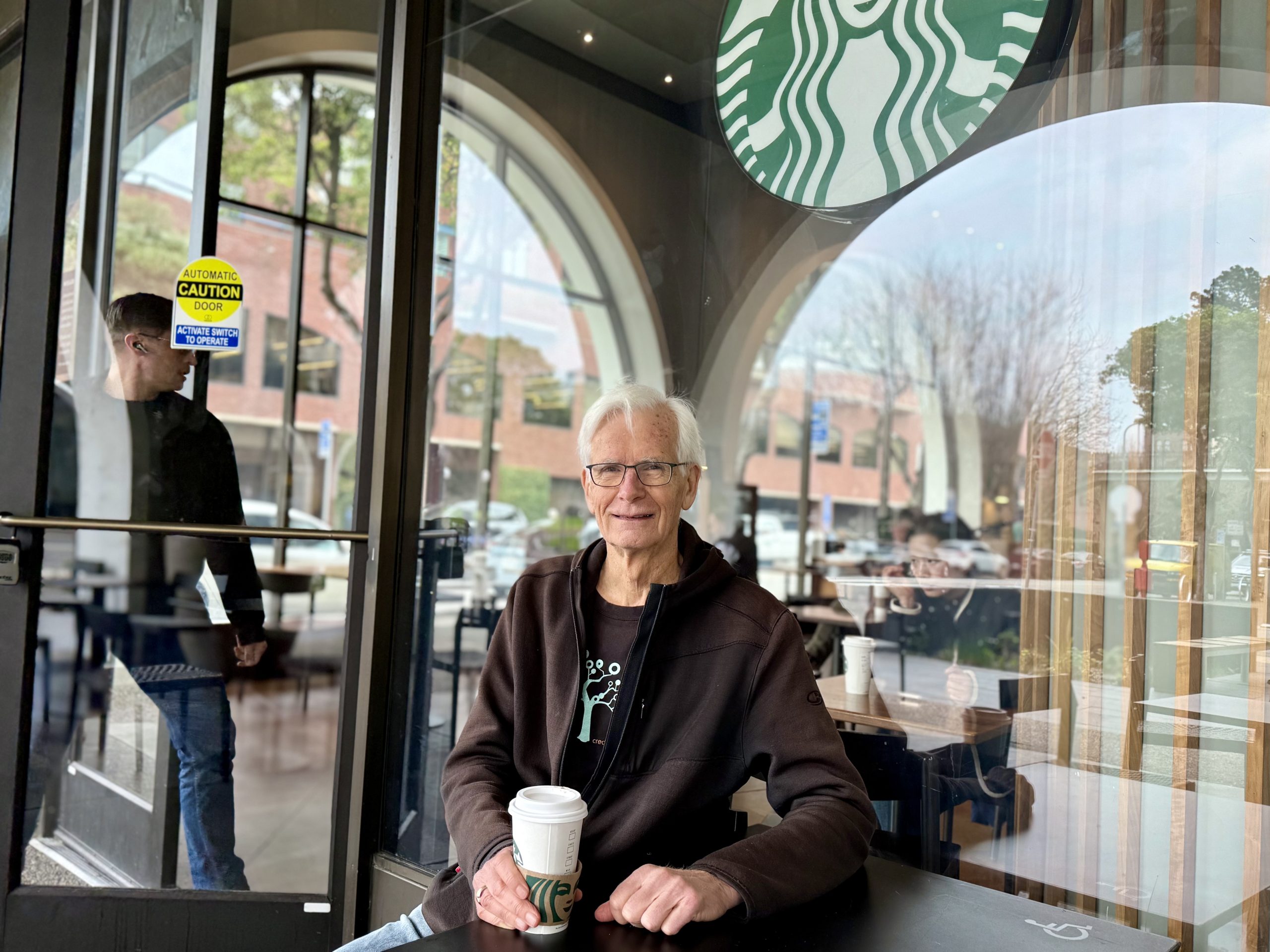 An elderly person holds a Starbucks cup, sitting outside a Starbucks cafe. Reflections in the glass show a street and another person entering.