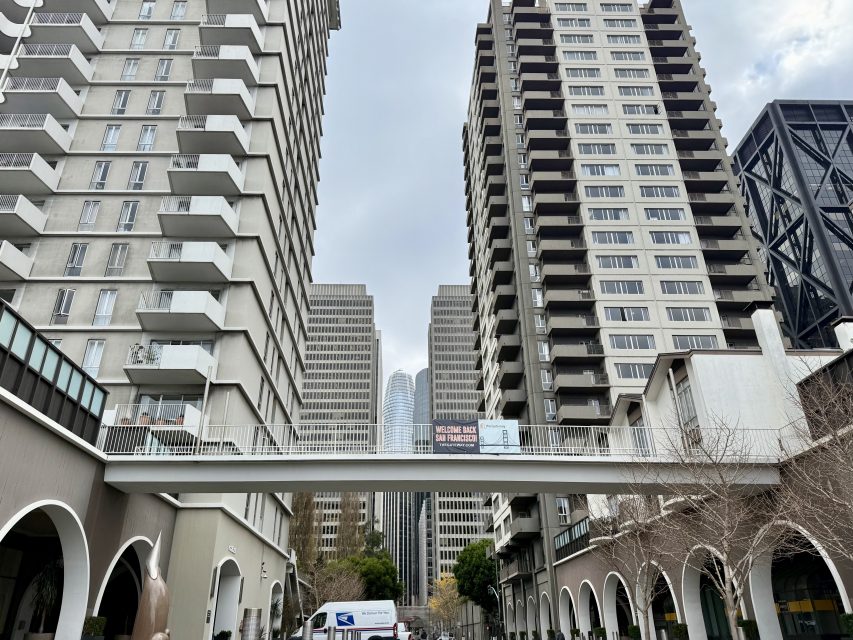 A view of a cityscape with tall buildings and a skybridge connecting two of them. A van and trees are visible on the street below. The sky is overcast.