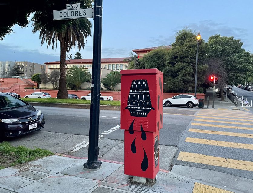 Red utility box with black and white abstract design, resembling a face with teeth, on a street corner near Dolores Street sign. Cars, trees, and buildings in the background.