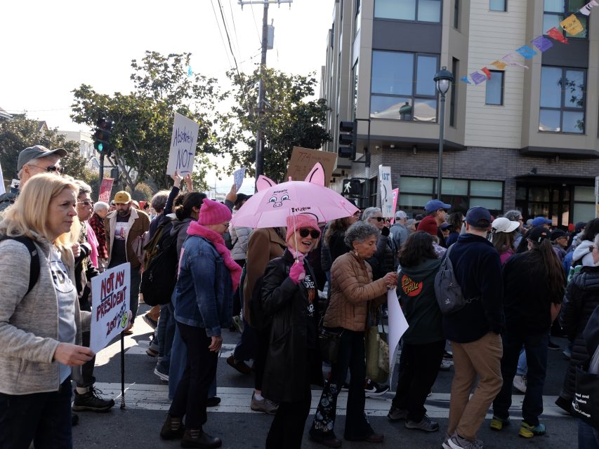 A group of people are participating in a street protest. Some are holding signs and a pink umbrella. They are gathered in front of a building with colorful flags above.