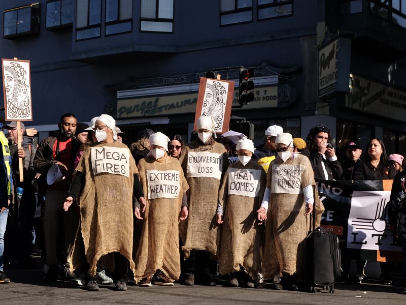 A group of people in burlap sack costumes and white masks protest climate issues with signs reading "Mega Fires," "Extreme Weather," and "Biodiversity Loss" on a city street.