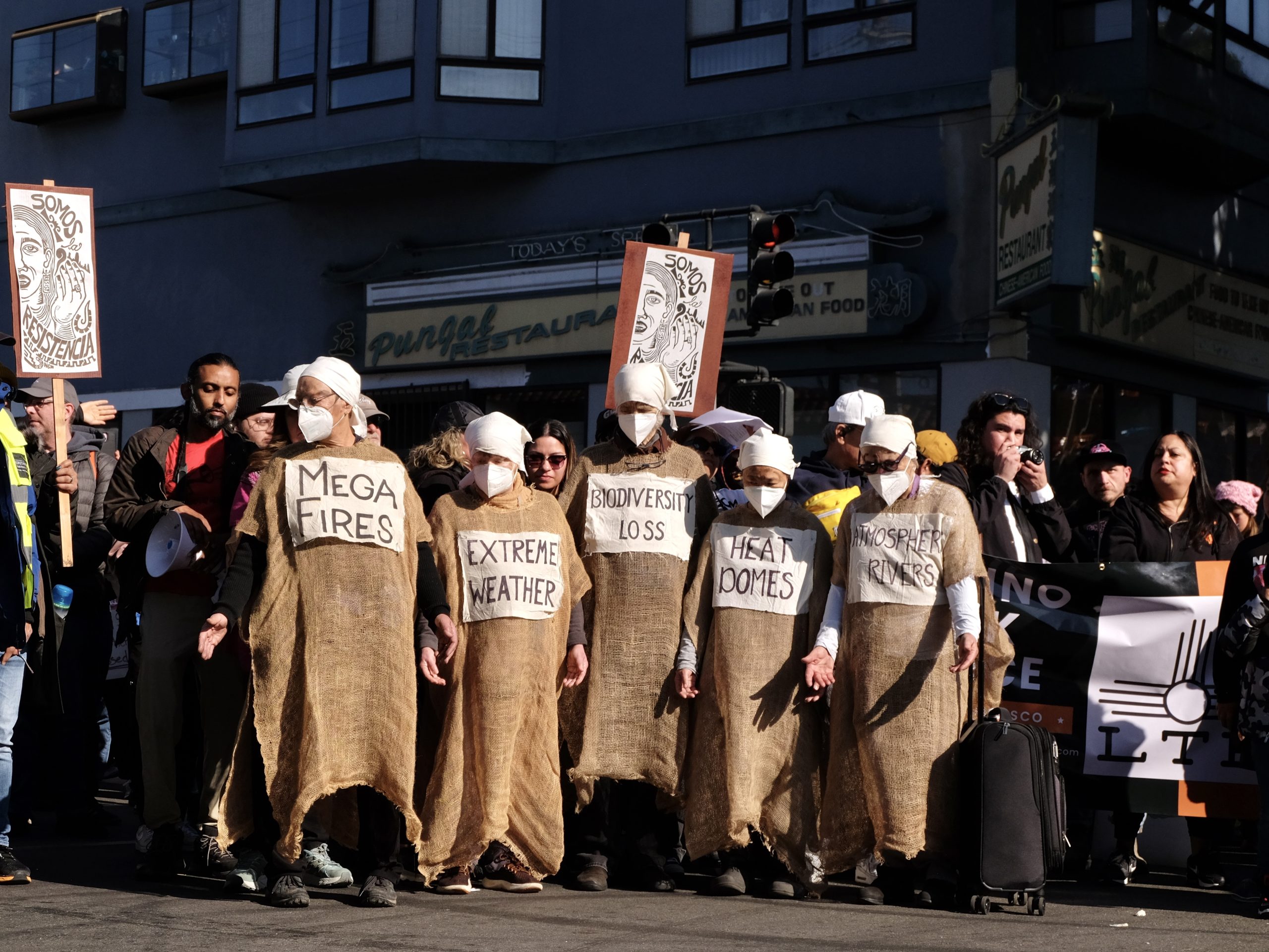 A group of people in burlap sack costumes and white masks protest climate issues with signs reading "Mega Fires," "Extreme Weather," and "Biodiversity Loss" on a city street.