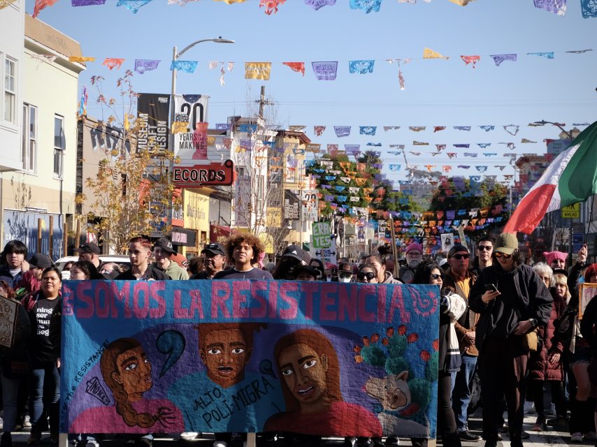A group of people march down a street holding a banner with "Somos La Resistencia" in a colorful setting with decorative flags overhead.