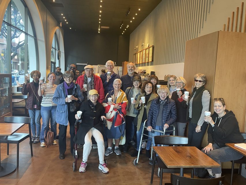 A group of people pose together inside a coffee shop, holding cups. Some are sitting, and others are standing, with tables and chairs around them.