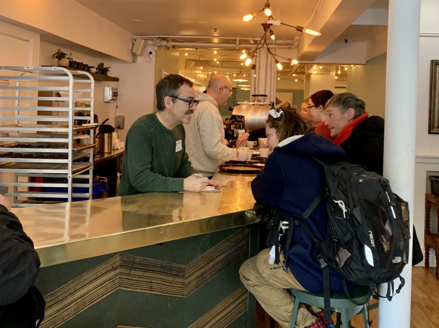 Customers are ordering at a cafe counter. A barista is speaking with a seated customer wearing a backpack. Other patrons are lined up and engaged in conversation.