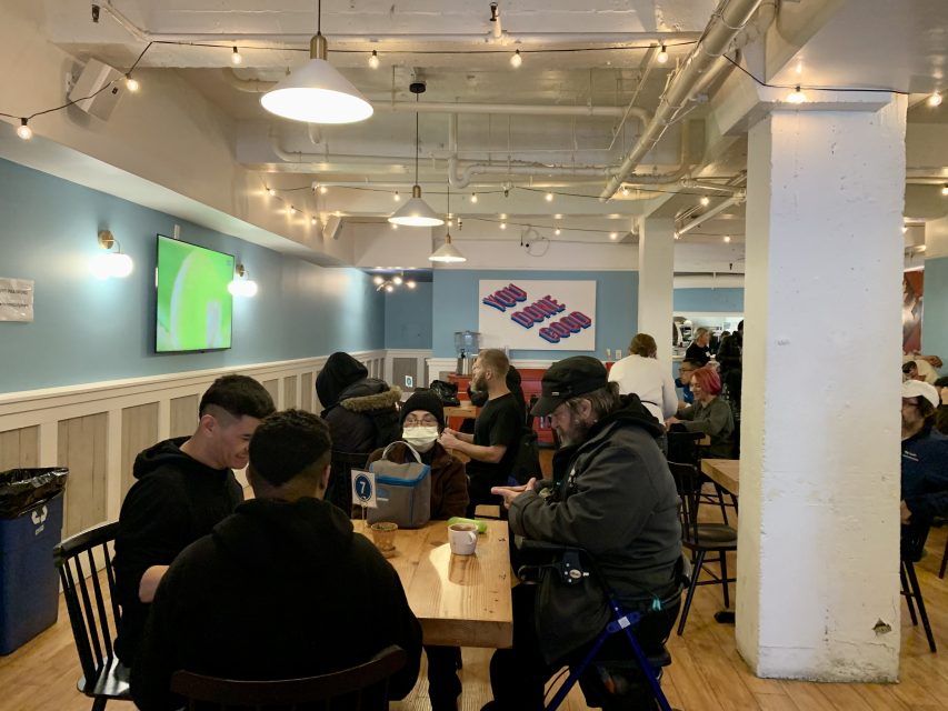 People sitting at wooden tables in a cafe with light blue walls and string lights. A sign on the wall says "YOU DONE GOOD." A TV displays a green screen.