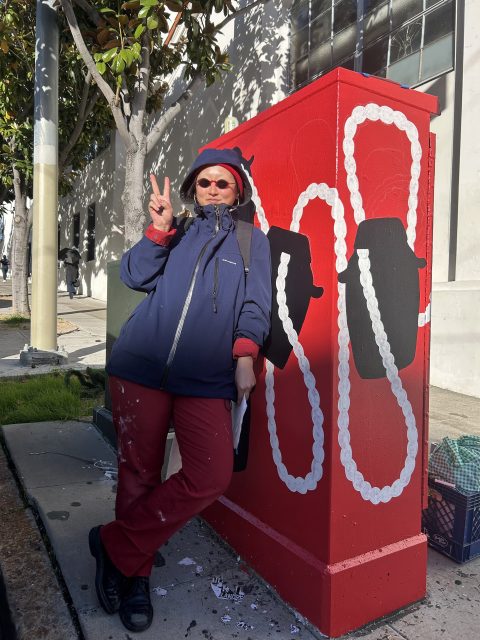 A person in a blue jacket and red pants stands on a sidewalk, posing with a peace sign next to a red-painted utility box featuring black shapes and white patterns.