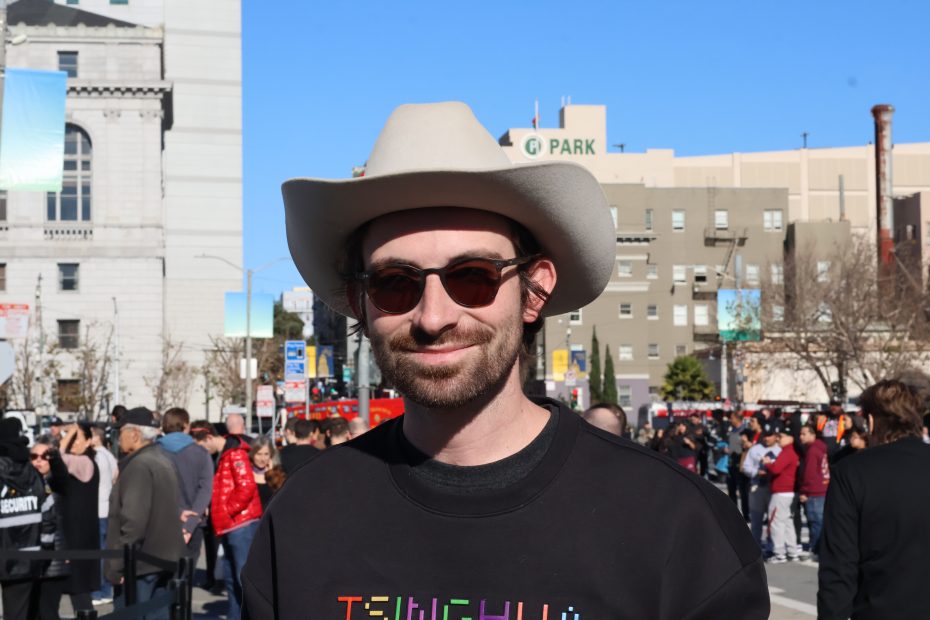 A person wearing sunglasses and a cowboy hat smiles in front of an urban backdrop with buildings and people around.