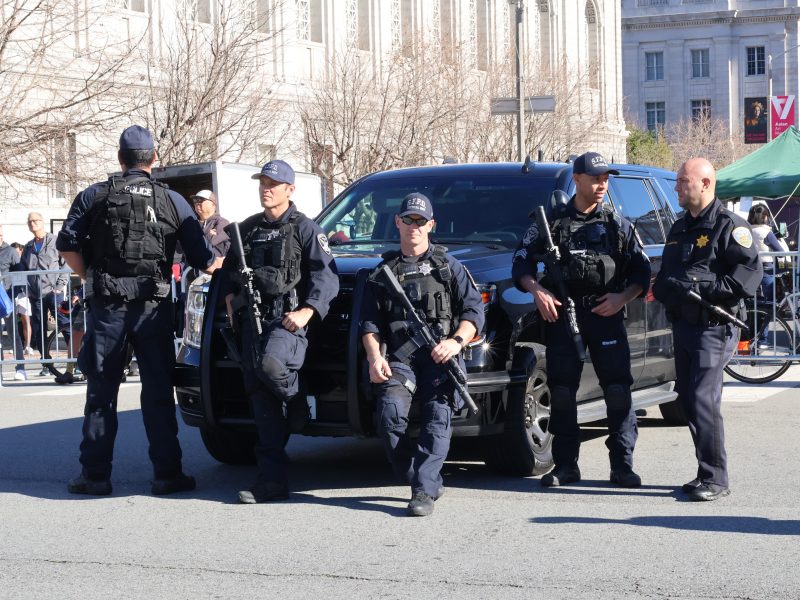 A group of police officers in tactical gear stand in front of a black SUV on a city street.