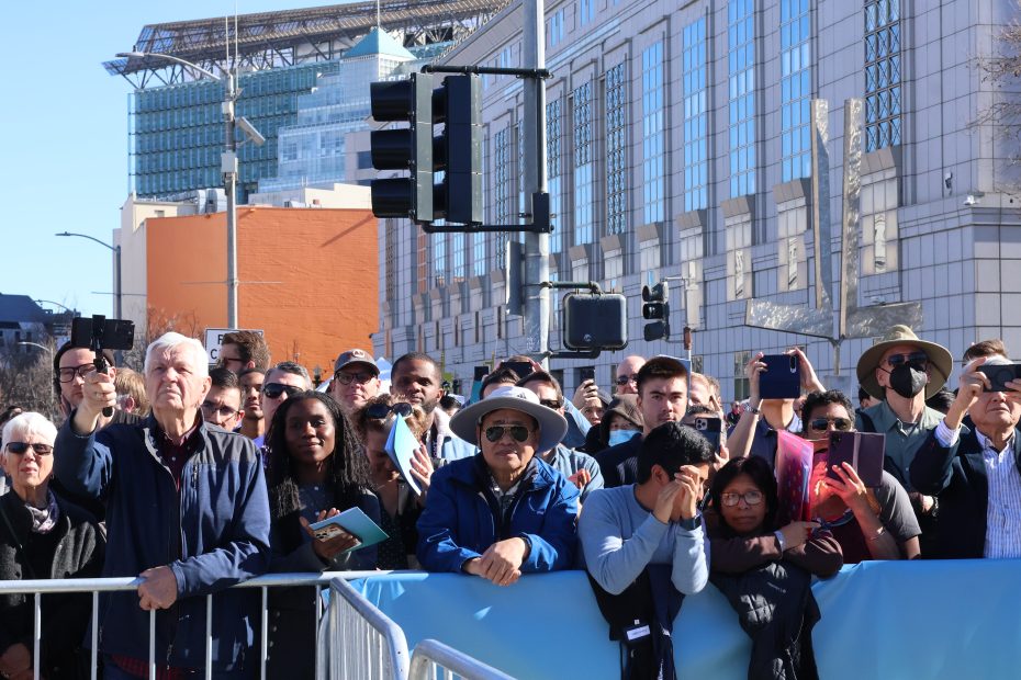 A diverse crowd stands behind a barricade on a sunny day, some holding phones up to take photos. A modern building is visible in the background.