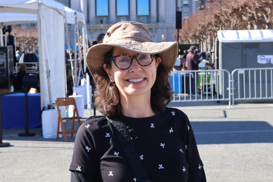 A person wearing a wide-brimmed hat and glasses smiles outdoors at an event.