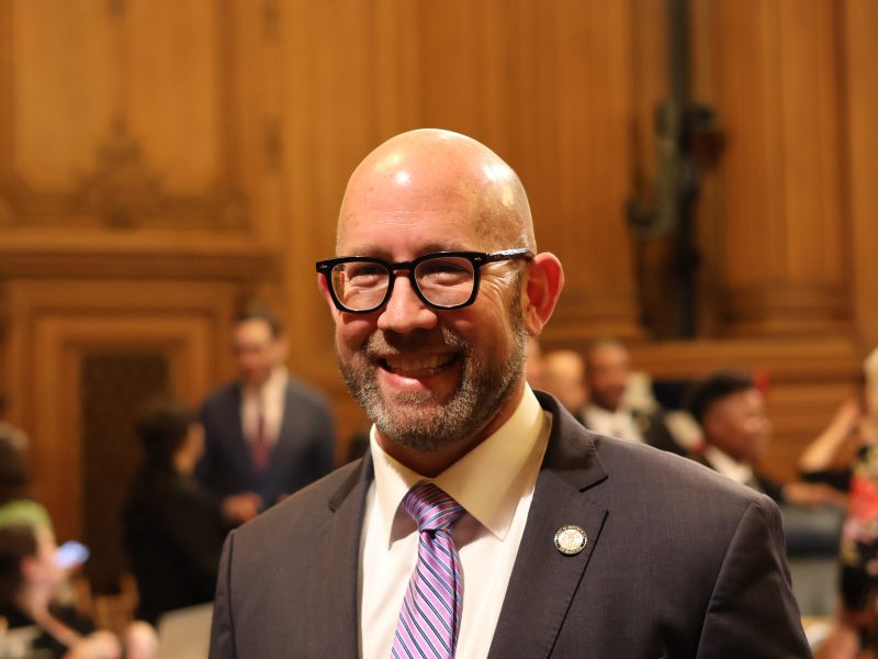 A man in a suit and tie smiles in a wood-paneled room with people in the background.
