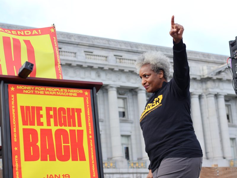 Person speaking at a rally, raising their right arm. Red and yellow signs read "WE FIGHT BACK." A classical building is in the background.