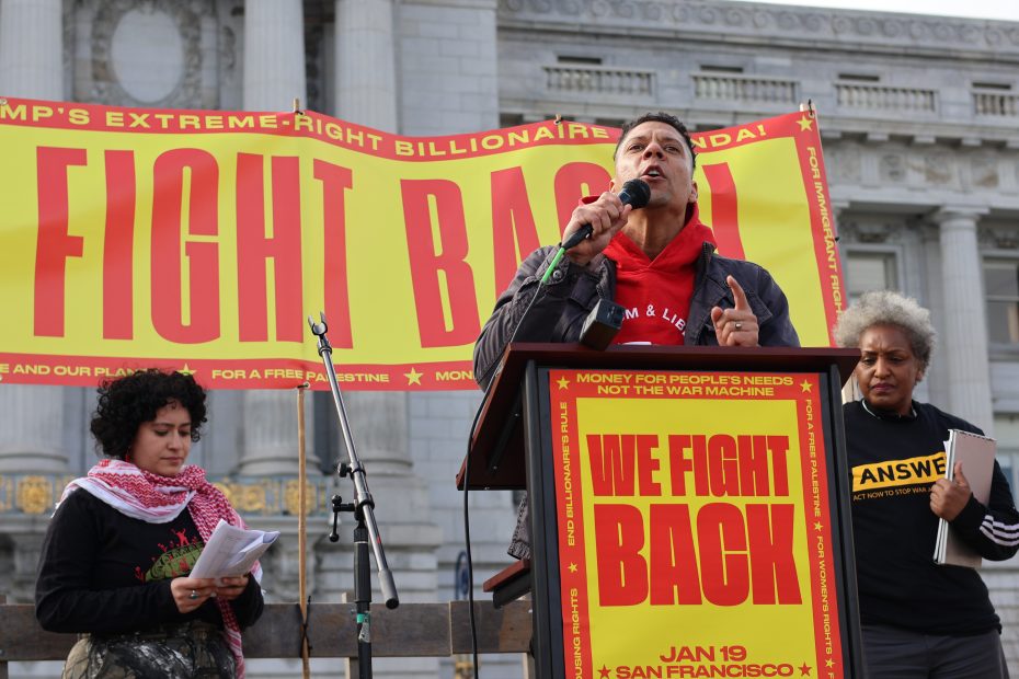 Person speaking at a rally in front of a banner that reads "Fight Back" and "We Fight Back," with two people standing nearby.