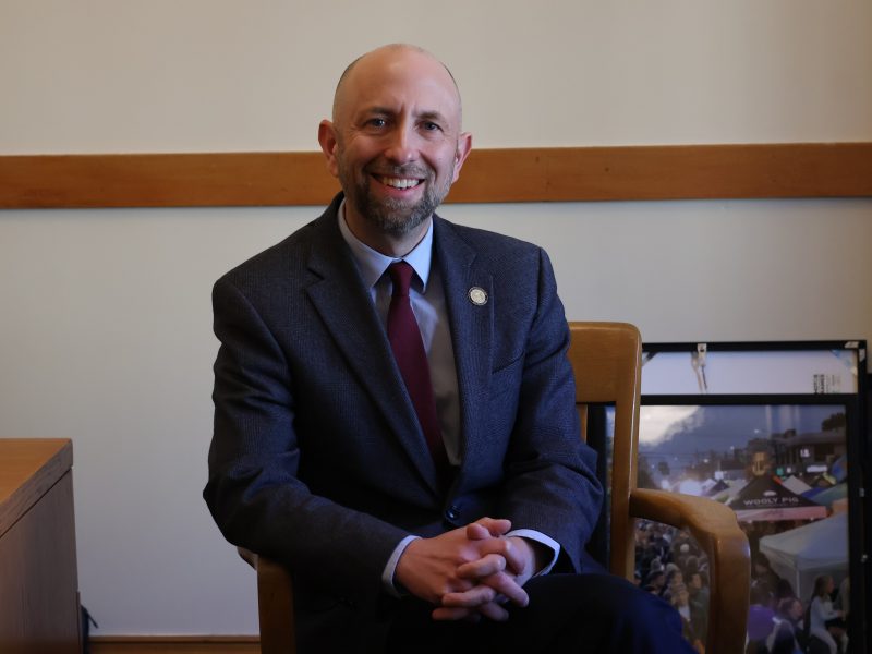 A man in a suit and tie sits in a wooden chair, smiling, with framed pictures in the background.