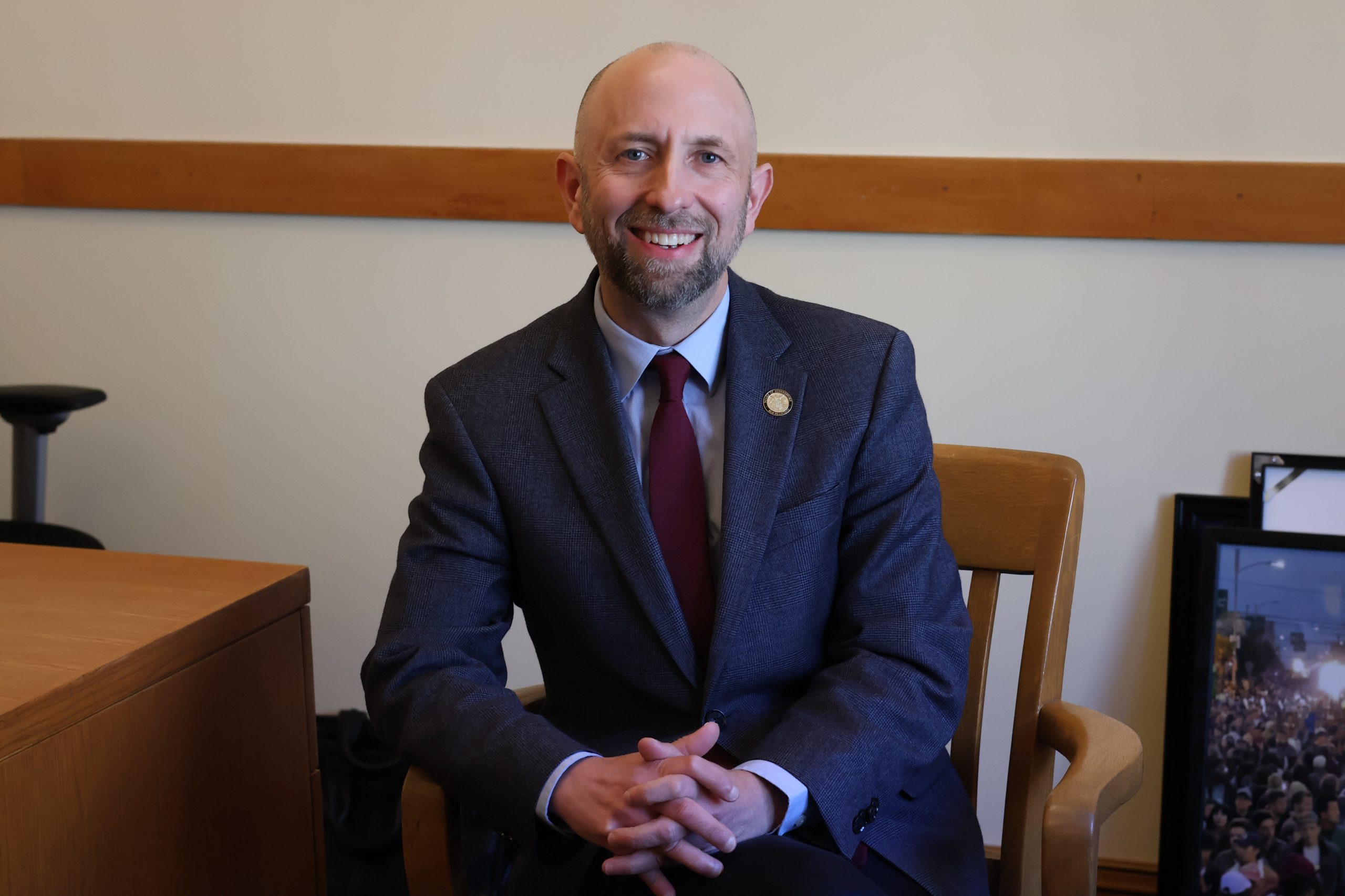 A man in a suit and tie sits on a wooden chair in an office setting.