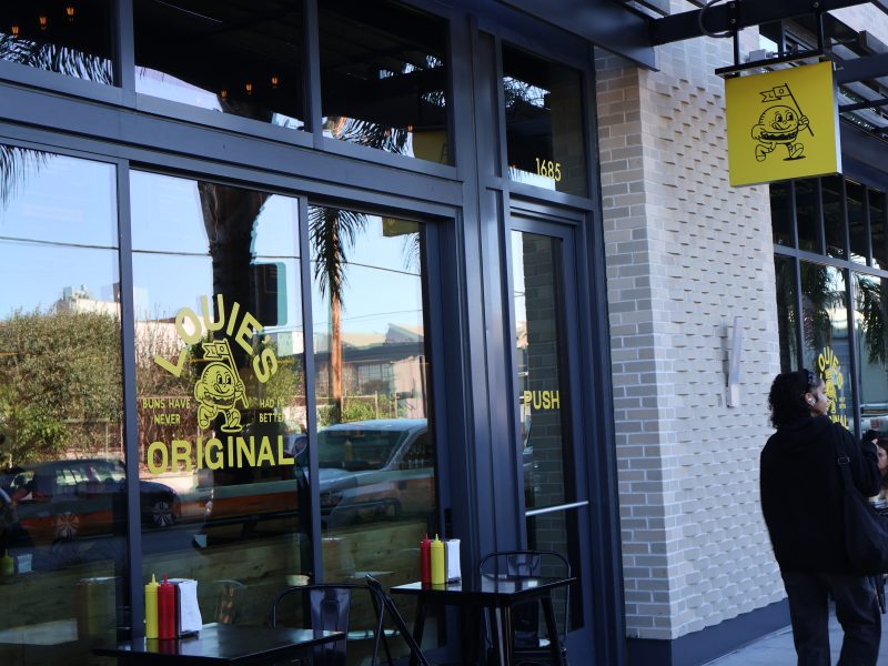 Street view of a restaurant facade with large windows, displaying yellow signage featuring a cartoon character. Tables with condiments are set up outside.