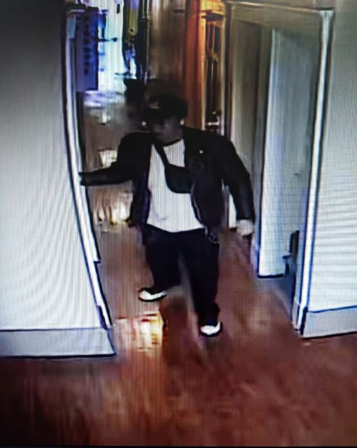 A person in a cap and jacket cautiously walks down a hallway with wooden floors, holding a door frame, amid whispers of a recent police shooting resonating through the air.