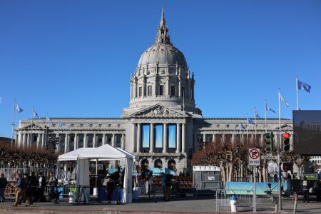 A large domed building with columns, serving as a hub for nonprofit initiatives, is fenced off with security tents and barricades under a clear blue sky.