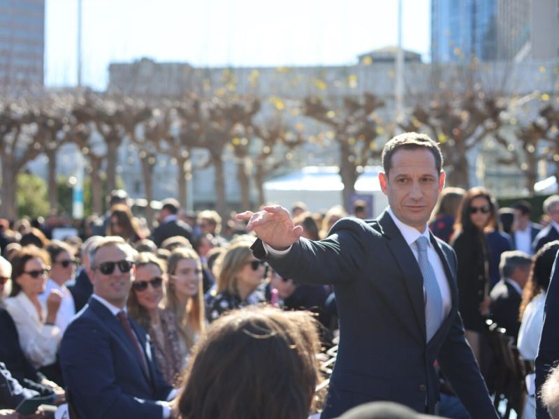 A man in a suit gestures in front of a seated crowd outdoors, with trees and city buildings in the background.