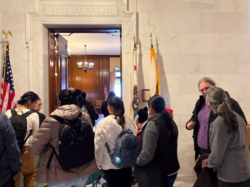 A group of people stand outside a room marked "The Mayor" in a building with marble walls and American flags.