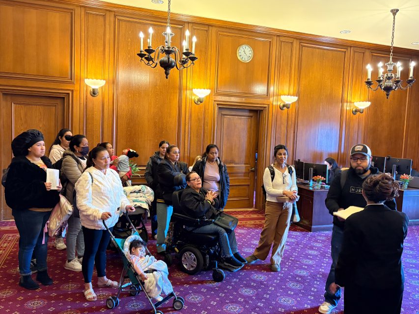 A group of people, including children in strollers and a person in a wheelchair, listen to a speaker in a wood-paneled room with chandeliers and a red carpet.