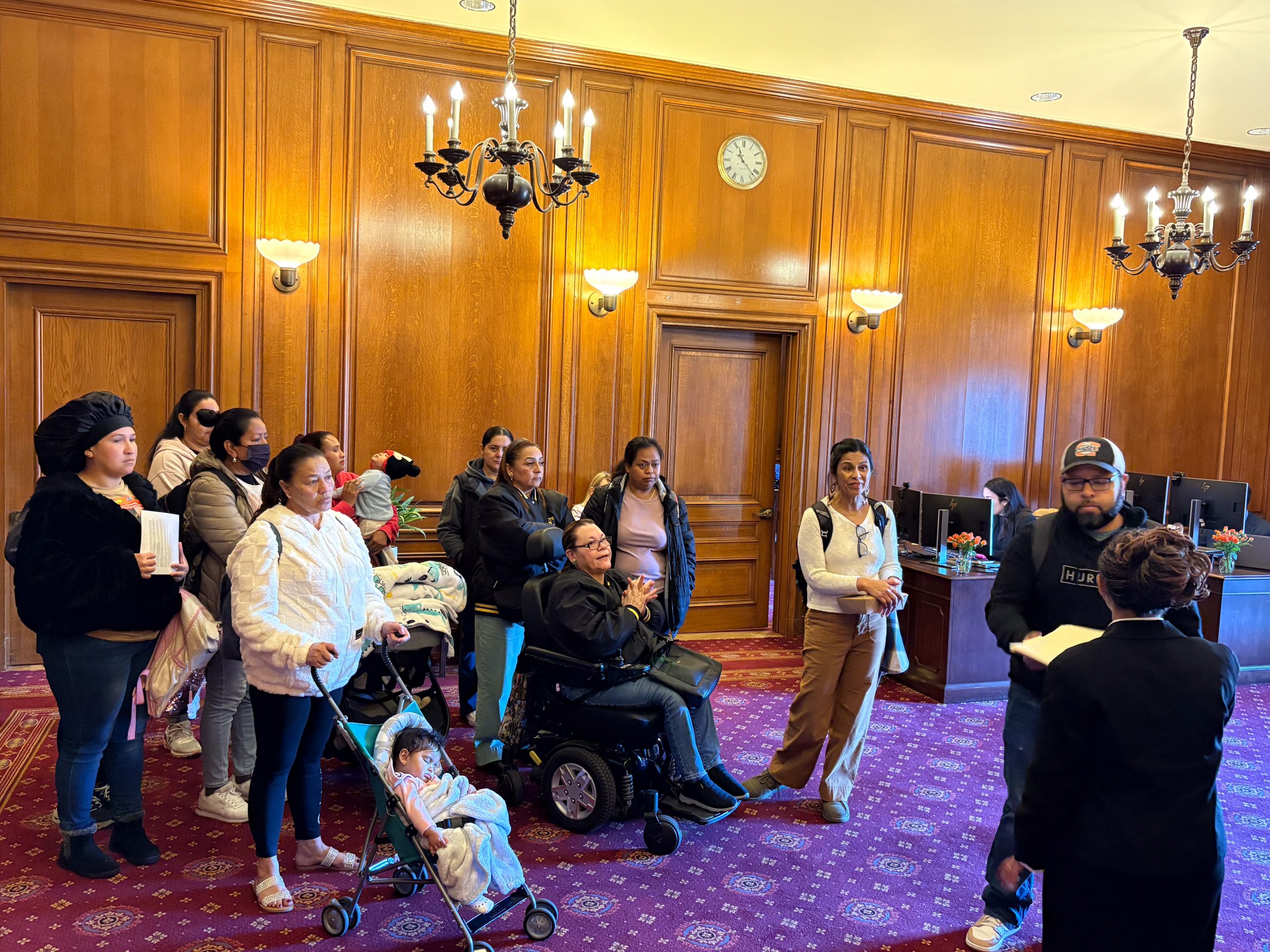 A group of people, including children in strollers and a person in a wheelchair, listen to a speaker in a wood-paneled room with chandeliers and a red carpet.