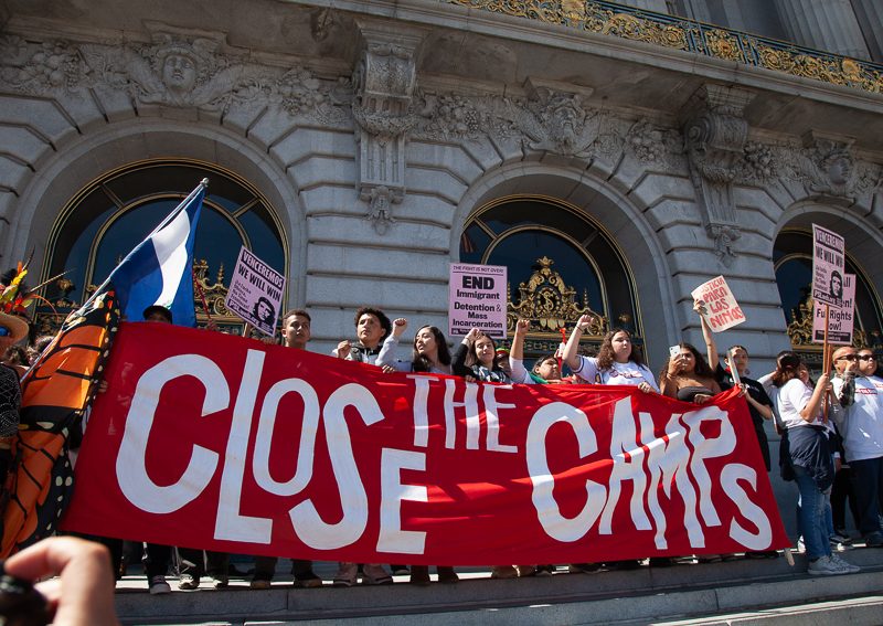 抗議者站在建築物台階上，手持一面巨大的 [Close the Camps] 橫幅，部分人還舉著較小的標語牌。支持移民權益。