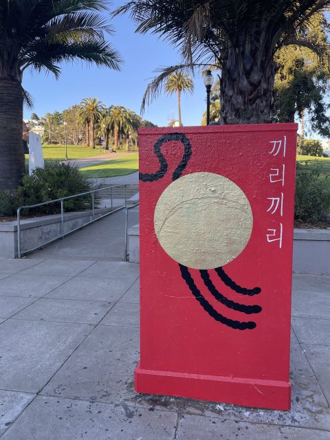 Red utility box with abstract design, featuring a yellow circle and black curved lines. Korean characters on the side. Palm trees and grass in the background.