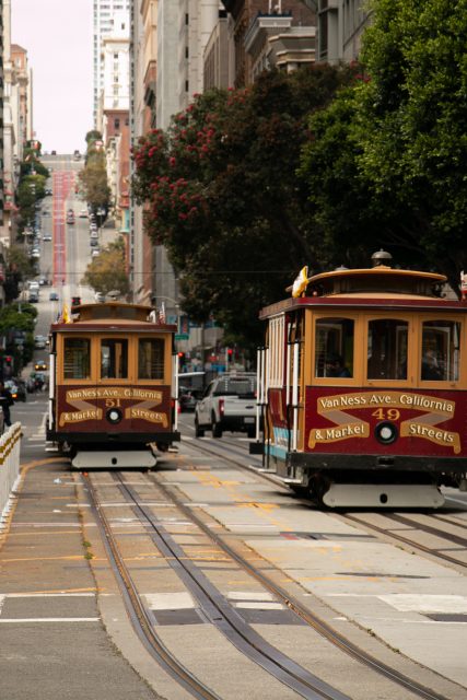 Two vintage cable cars travel along a street with tracks in a city, surrounded by tall buildings and trees.