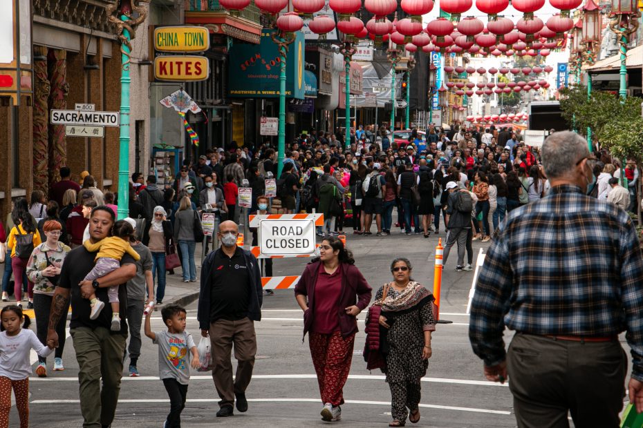 A busy street in Chinatown with crowds of people walking, red lanterns overhead, and a "Road Closed" sign in the background.