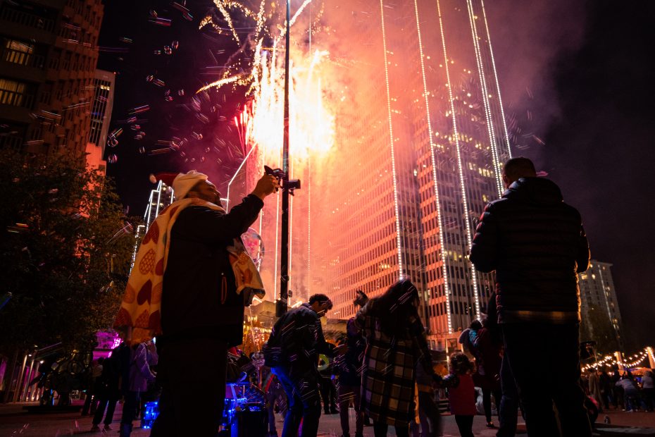 People watching fireworks near tall buildings at night, with some capturing the moment on their phones.
