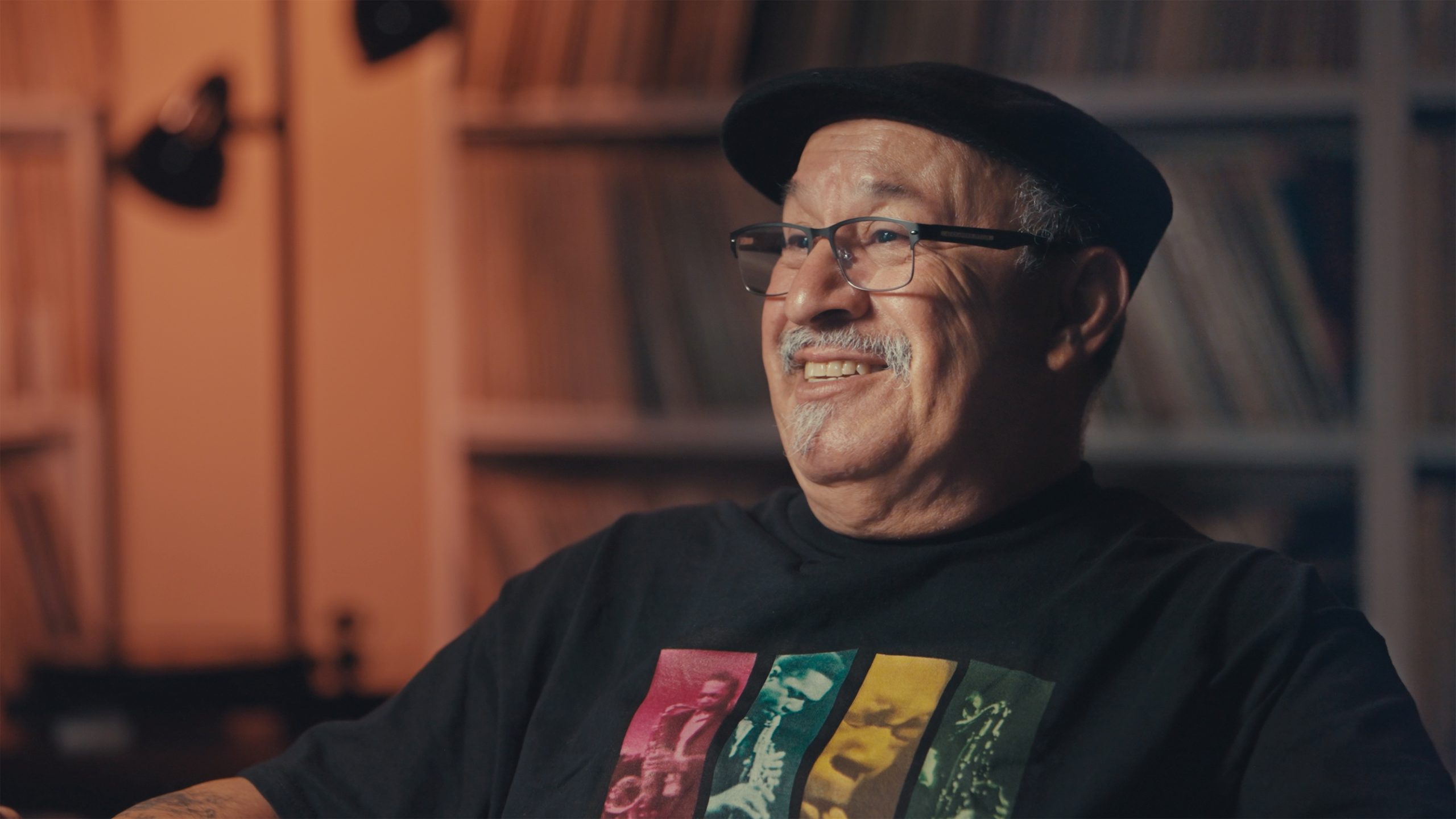 Smiling man with a mustache and glasses, wearing a black cap and T-shirt, sits in front of blurred shelves.