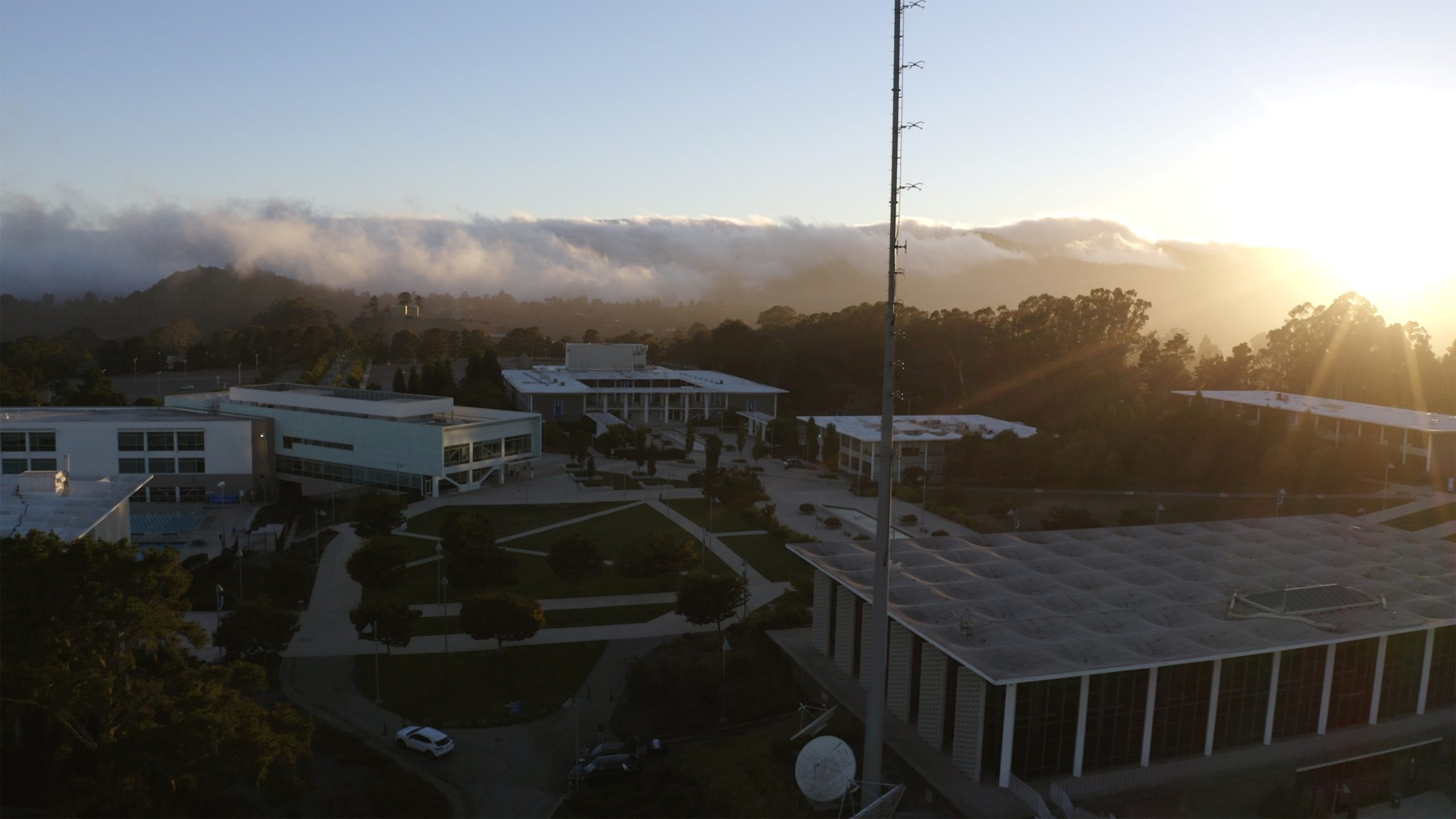 Aerial view of a university campus at sunset with modern buildings, trees, and nearby hills partially shrouded in mist.