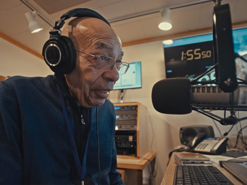 Elderly man in a radio studio wearing headphones and speaking into a microphone, surrounded by equipment and screens.