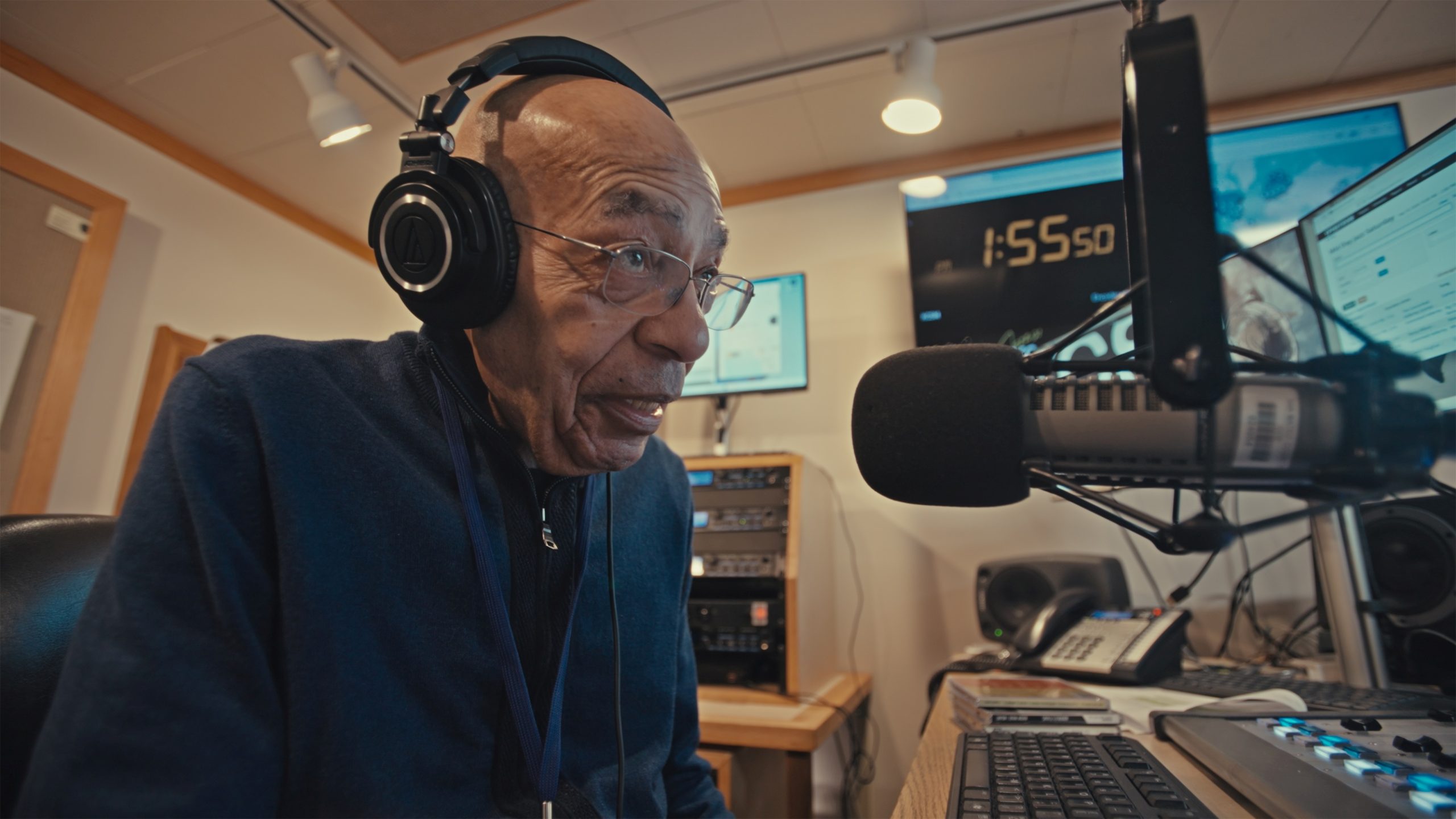 Elderly man in a radio studio wearing headphones and speaking into a microphone, surrounded by equipment and screens.