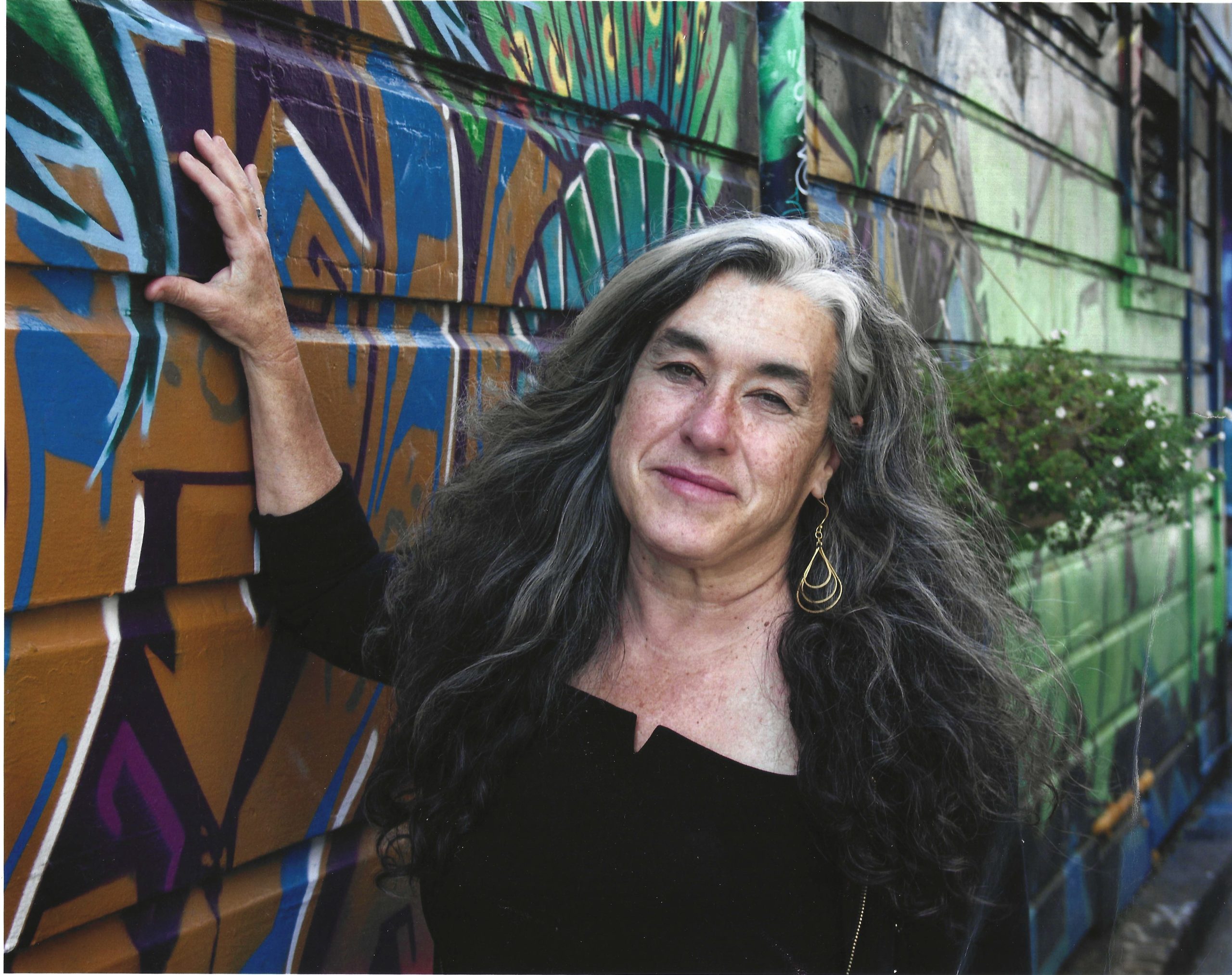 A woman with long gray hair leans against a colorful graffiti-covered wall, wearing a black top and gold earrings.