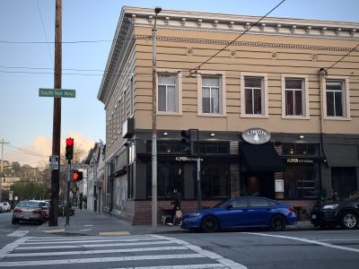 Street corner with a three-story building housing "Limon Rotisserie." A blue car is parked outside. A traffic light and "South Van Ness" street sign are visible.