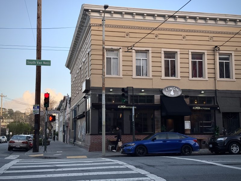 Street corner with a three-story building housing "Limon Rotisserie." A blue car is parked outside. A traffic light and "South Van Ness" street sign are visible.
