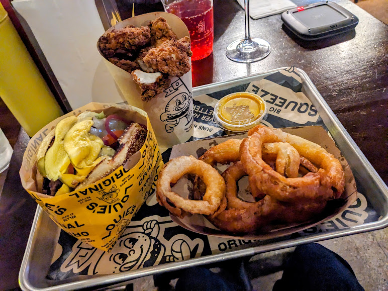 A tray with a cheeseburger, onion rings, fried chicken, dipping sauce, a glass of red drink, and a yellow bottle, on a dark table.